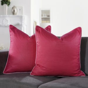 May include: Two magenta throw pillows with white rope trim are displayed on a gray sofa. The pillows have a textured appearance and a classic, tailored design. The pillows add a pop of color to the neutral-toned living room.