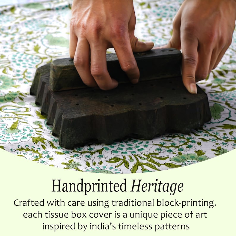 Close-up of hands using a traditional block printing tool on fabric with a green floral pattern. Text below reads: "Handprinted Heritage - Crafted with care using traditional block-printing. Each tissue box cover is a unique piece of art inspired by India’s timeless patterns."