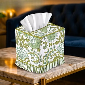 Op de afbeelding: Een vierkante tissueboxhoes met een wit en groen bloemenpatroon. De hoes heeft een groene rand en een gefranjerde rand. Witte tissues zijn zichtbaar aan de bovenkant. De doos staat op een marmeren tafel.