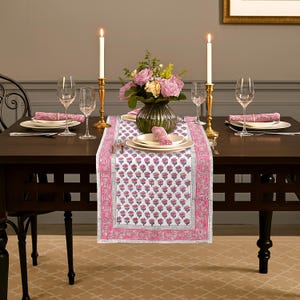 May include: A dining table set for a meal, featuring a white and pink floral patterned table runner. The table is set with plates, glasses, and napkins. Two lit candles in gold holders flank a vase of pink flowers.