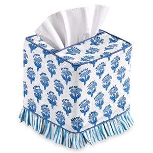 Op de afbeelding: Een vierkante tissueboxhoes met een witte achtergrond en een blauw bloemenpatroon. De boven- en randen zijn afgewerkt met blauwe biezen. De onderkant heeft een blauw-wit gestreepte ruche. Witte tissues zijn zichtbaar aan de bovenkant.