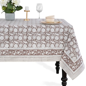 Puede incluir: Un mantel rectangular con un estampado floral marrón y blanco. El mantel está sobre una mesa de madera oscura. El diseño floral incluye formas circulares y de hojas. El borde presenta un patrón de rayas.