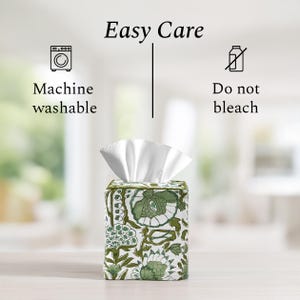 Fabric tissue box cover with green floral design on a light wood surface with blurred background. Text reads: "Easy Care" with icons and instructions: "Machine washable" and "Do not bleach."