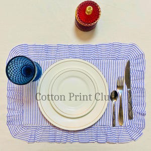 Puede incluir: Una mesa puesta con un mantel individual a rayas azules y blancas, dos platos blancos con ribete dorado y un vaso azul. Cubiertos y un tarro rojo con tapa dorada también están presentes. El texto "Cotton Print Club" es visible.
