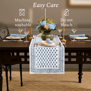 Elegant dining table setup featuring a blue and white patterned table runner, matching napkins, and a floral centerpiece with blue roses. Surrounded by white plates, wine glasses, and gold-toned candle holders. The image highlights care instructions: "Machine washable" and "Do not bleach.