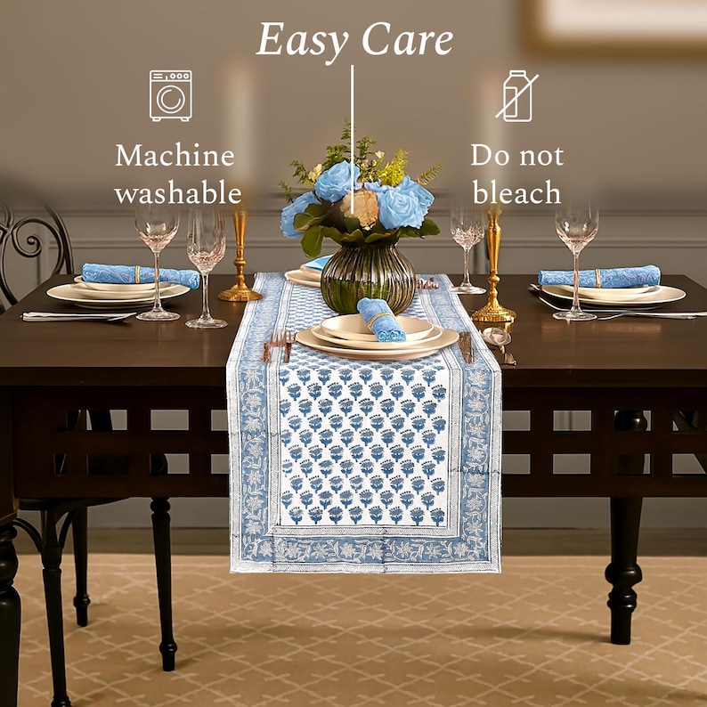 Elegant dining table setup featuring a blue and white patterned table runner, matching napkins, and a floral centerpiece with blue roses. Surrounded by white plates, wine glasses, and gold-toned candle holders. The image highlights care instructions: "Machine washable" and "Do not bleach.