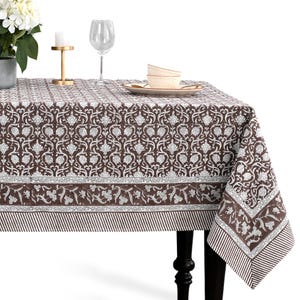 Puede incluir: Un mantel con estampado floral marrón y blanco. El mantel presenta un diseño detallado con un borde de patrones florales y a rayas. La mesa está puesta con una copa de vino, platos y un candelabro.