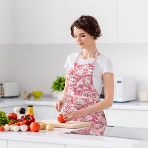 May include: A red and white floral print apron with a striped pocket and neck strap. The apron is being worn in a kitchen setting, with fresh vegetables and a cutting board visible on the counter.