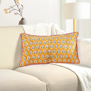 May include: A rectangular decorative pillow with a mustard yellow background and a repeating floral pattern in white and pink. The pillow has a pink border and is placed on a cream-colored sofa.