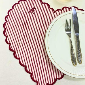 Puede incluir: Un mantel individual en forma de corazón con borde festoneado, con rayas verticales rojas y blancas. Un plato blanco con borde dorado, un tenedor y un cuchillo están colocados sobre el mantel individual. El mantel individual tiene la letra "A".