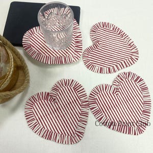 Op de afbeelding: Hartvormige onderzetters met een rood-wit gestreept patroon en gerimpelde randen. Een helder glas staat op een van de onderzetters. De stoffen onderzetters zijn ontworpen om oppervlakken te beschermen.