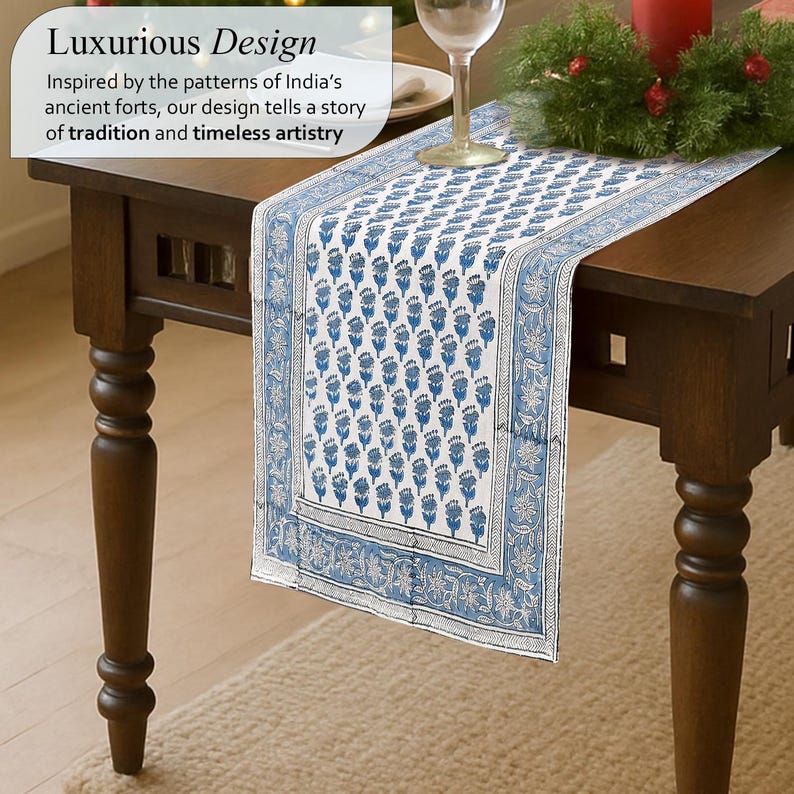 Luxurious Apatite blue table runner with intricate floral patterns inspired by India’s ancient forts, displayed on a dark wooden table. Text overlay highlights the design's roots in tradition and timeless artistry, suggesting elegance and cultural richness. A wine glass and festive greenery add a sophisticated touch to the scene.