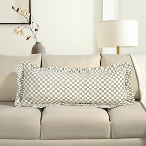 May include: A rectangular decorative pillow with a checkered pattern in shades of green and white, framed by a ruffled edge. The pillow is placed on a beige sofa, suggesting a home decor theme.