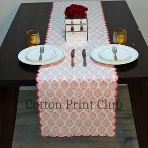 Könnte beinhalten: Eine Tischdekoration mit einem weiß-roten Tischläufer mit gewelltem Rand. Der Tisch ist mit weißen Tellern, Besteck und goldfarbenen Kerzenhaltern gedeckt. Eine verspiegelte Box und rote Blumen befinden sich in der Mitte.