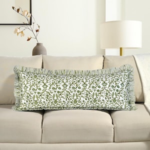 Pode incluir: Uma almofada retangular decorativa com fundo branco e padrão floral verde. A almofada tem uma borda com babados com listras verdes e brancas. A almofada é colocada em um sofá bege.