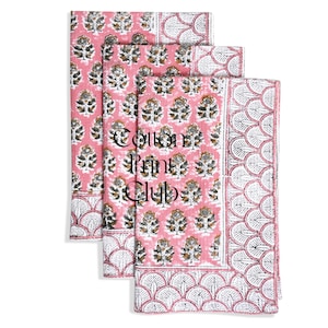May include: A stack of pink cotton fabric with a floral pattern and white scalloped border. The fabric has a repeating pattern of stylized flowers in shades of grey and gold. The text "Cotton Print Club" is printed on the fabric.