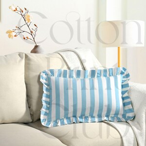 Può includere: Un cuscino rettangolare con un motivo a righe blu e bianche e un bordo a balze. Il cuscino è posto su un divano beige, con altri cuscini e una coperta visibili sullo sfondo. Una lampada e un vaso con fiori sono anch'essi sullo sfondo.