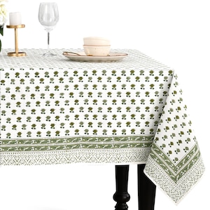Puede incluir: Mantel blanco con un patrón repetido de diseños florales verdes. El mantel tiene un borde decorativo con un patrón detallado en verde y blanco. Un plato, cuencos y una copa de vino están sobre la mesa.
