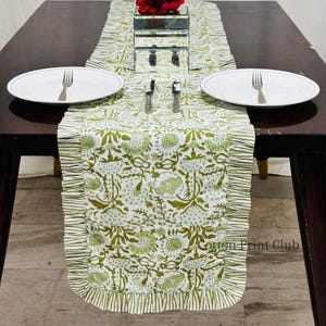 May include: A white and green floral table runner with a ruffled edge, set on a dark wood table. Two white plates with forks and two knives are placed on the table. The text "Cotton Print Club" is visible on the runner.
