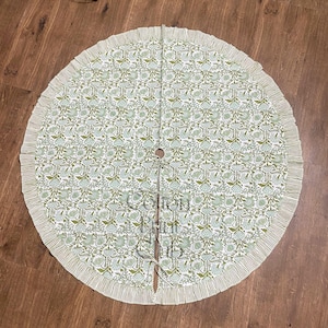 May include: A round Christmas tree skirt with a floral pattern in shades of green and white. The skirt has a ruffled edge and a split opening with ties. The overall design is a classic, elegant style.