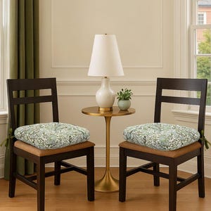 May include: Two dark brown wooden chairs with floral patterned seat cushions. A gold side table with a white lamp and a small potted plant are between the chairs. The chairs are in front of a window.