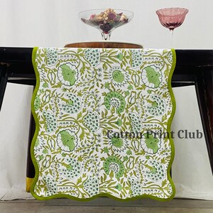 May include: A white table runner featuring a repeating green floral pattern with scalloped edges trimmed in solid green. The fabric displays the text Cotton Print Club in black lettering. The runner is draped over a dark surface with glassware.