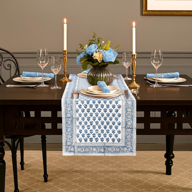 May include: A dining table set for a meal, featuring a blue and white floral patterned table runner. The table is set with plates, napkins, silverware, and wine glasses. Two lit candles in gold candle holders flank a vase of blue flowers.