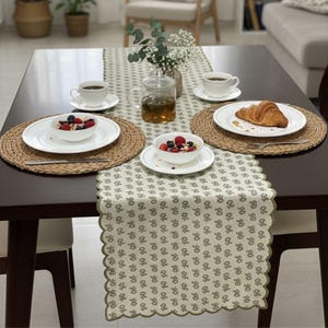 Puede incluir: Una mesa de comedor de madera marrón oscuro preparada para el desayuno. La mesa está puesta con platos blancos, cuencos de bayas, tazas de café, una tetera y un cruasán. Los manteles individuales tejidos y un camino de mesa estampado completan la decoración.
