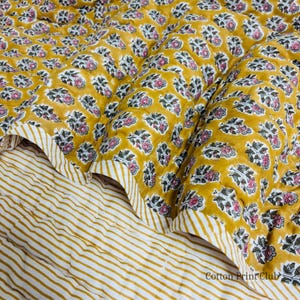 May include: A quilted blanket with a floral pattern in shades of yellow, pink, and gray. The blanket has a striped border and a striped underside. The text "Cotton Print Club" is visible in the bottom right corner.