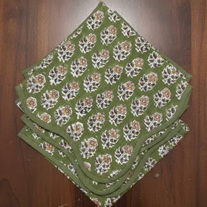 Puede incluir: Una pila de cuatro servilletas cuadradas de tela verde. Cada servilleta presenta un patrón floral repetido en blanco, marrón y melocotón. Los bordes están acabados con un ribete verde a juego. Las servilletas están apiladas sobre una superficie de madera.