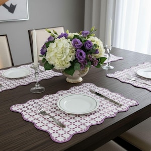 Può includere: Un tavolo da pranzo apparecchiato per un pasto. Il tavolo è apparecchiato con piatti bianchi, tovagliette fantasia con bordo viola, posate e un centrotavola floreale con fiori bianchi e viola. Due candele bianche in portacandele di vetro.