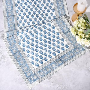 Hand block printed cotton tablecloth in blue and white floral design with intricate border. Apatite blue botanical motifs arranged in repeat pattern on white background, bordered with detailed floral vines. Perfect for dining table, coffee table, console, or outdoor patio décor. Artisan-made textile displayed with flowers and candle for elegant home styling.