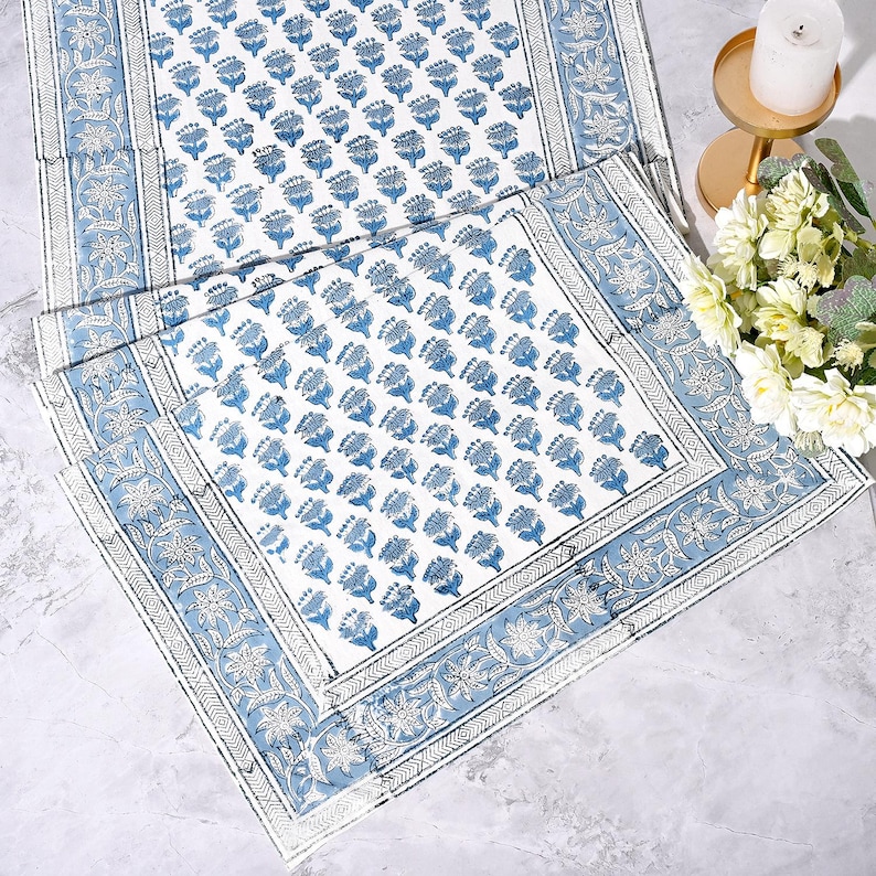 Hand block printed cotton tablecloth in blue and white floral design with intricate border. Apatite blue botanical motifs arranged in repeat pattern on white background, bordered with detailed floral vines. Perfect for dining table, coffee table, console, or outdoor patio décor. Artisan-made textile displayed with flowers and candle for elegant home styling.