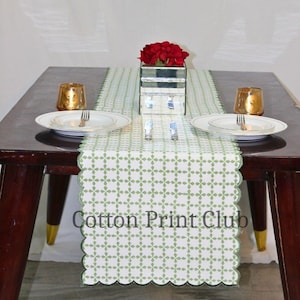 Puede incluir: Una mesa de comedor preparada para una comida, con un camino de mesa con estampado blanco y verde con bordes festoneados. La mesa está puesta con platos blancos, vasos con borde dorado, cubiertos y un jarrón espejado con flores rojas. El texto "Cotton Print Club" es visible.