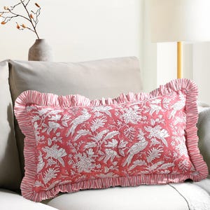 Puede incluir: Un cojín rectangular decorativo con un estampado floral y de pájaros en color rosa. El cojín tiene un borde con volantes y está colocado sobre un sofá de color claro. El diseño presenta pájaros y flores blancas sobre fondo rosa.