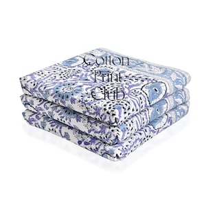 May include: A stack of three folded cotton fabric items with a floral pattern in shades of blue, purple, and white. The text "Cotton Print Club" is printed on the top item.