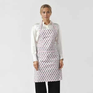 Peut inclure: Un tablier blanc avec un motif floral répétitif rose et bleu. Le tablier a une bordure et des bretelles rayées rouges et blanches. La personne sur l'image porte une chemise blanche boutonnée et un pantalon noir.