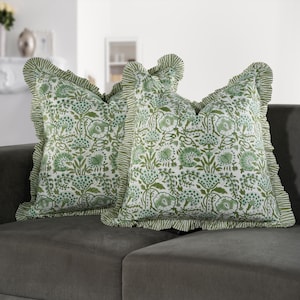 May include: Two decorative throw pillows with a white background and a green floral pattern. The pillows have ruffled edges with a striped design. The pillows are resting on a gray sofa.