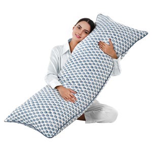 Può includere: Un lungo cuscino per il corpo rettangolare con sfondo bianco e motivo blu e bianco. Il cuscino ha un bordo blu scuro. Una persona con una camicia e pantaloni bianchi tiene il cuscino.