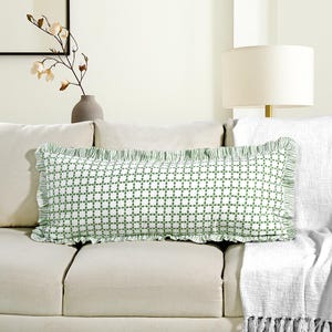 Puede incluir: Un cojín decorativo rectangular con fondo blanco y estampado floral verde. El cojín tiene un borde con volantes y está colocado sobre un sofá beige. El cojín mide aproximadamente 30 cm por 61 cm.