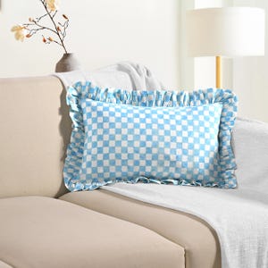Peut inclure: Un coussin rectangulaire à motif de carreaux bleus et blancs et bordure à volants. Le coussin est posé sur un canapé beige, avec une couverture blanche drapée sur le dossier.