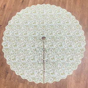 May include: A round Christmas tree skirt with a scalloped edge. The skirt features a repeating floral pattern in shades of green and white. It has a center opening and ties for securing around a tree trunk. The skirt is made of fabric.