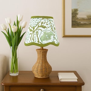 Puede incluir: Una lámpara de mesa decorativa con una base de mimbre tejido y una pantalla con estampado floral. La pantalla tiene un fondo blanco con diseños botánicos verdes y un ribete verde. La lámpara está sobre una mesita de noche de madera.