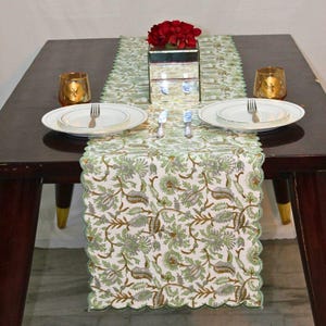 May include: A dining table set for a meal, featuring a floral patterned table runner in green and brown. White plates, silverware, and gold candle holders are arranged on the table. A mirrored vase holds red flowers.