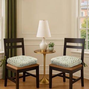 May include: Two dark brown wooden chairs with patterned seat cushions. The cushions have a white background with a green and light green leaf pattern. A small gold side table with a white lamp and a small plant sits between the chairs.