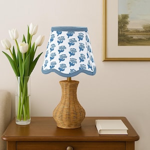 Peut inclure: Une lampe de table décorative avec un pied en osier tressé et un abat-jour blanc orné d'un motif floral bleu. L'abat-jour a un bord festonné et une bordure bleue. Un vase de tulipes blanches est à gauche de la lampe.