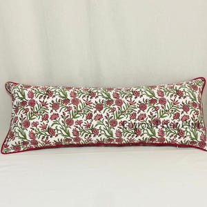 Puede incluir: Un cojín rectangular con un estampado floral rosa y verde sobre fondo blanco. El cojín tiene un borde rojo.