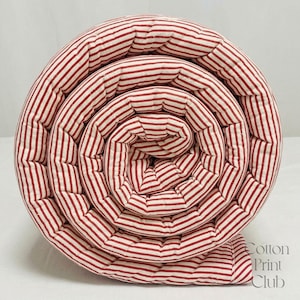 May include: A rolled textile featuring a repeating pattern of thin red stripes on a white background. The fabric is quilted with visible stitching lines running parallel to the stripes. The text Cotton Print Club is printed in the bottom right corner.
