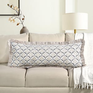 May include: A rectangular decorative pillow with a white background and a repeating pattern of stylized floral shapes in shades of gray and beige. The pillow has a ruffled edge with a striped border and is placed on a beige sofa.