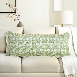 Könnte beinhalten: Ein rechteckiges Zierkissen mit einem grün-weißen Blumenmuster. Das Kissen hat einen gerüschten Rand und liegt auf einem weißen Sofa. Das Kissen ist etwa 76 cm lang.